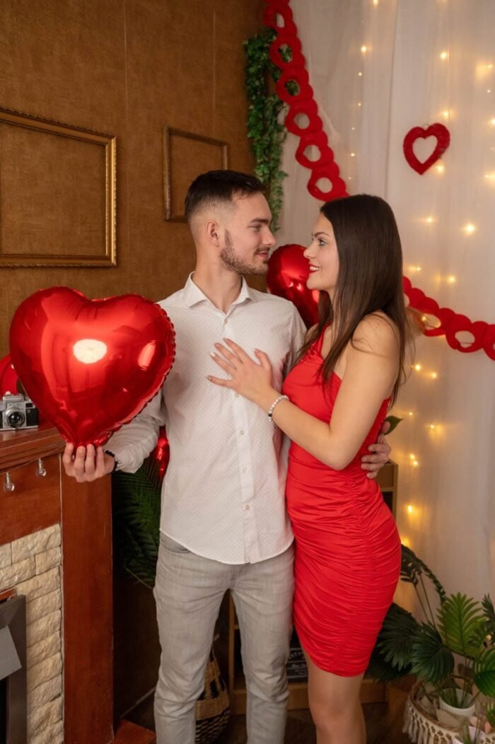 Fiatal pár romantikus Valentin-napi stúdiófotón, piros szív alakú lufival, meleg fények és dekorációk között.