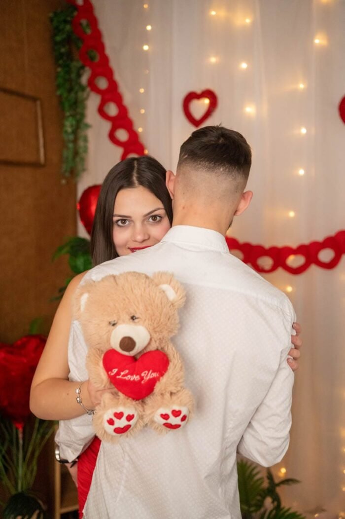 Fiatal nő öleli párját Valentin-napi stúdiófotón, kezében "I Love You" feliratú plüss macival, romantikus dekorációk előtt.