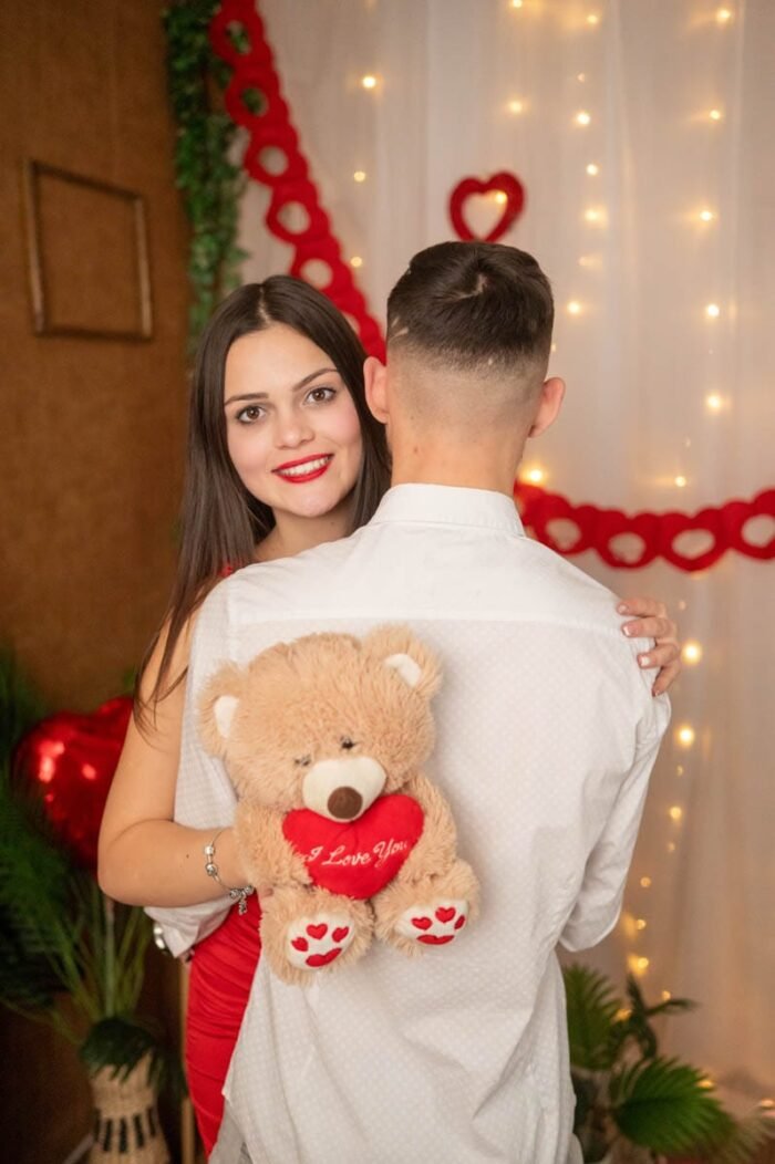 Fiatal nő mosolyog, miközben párját öleli Valentin-napi dekoráció előtt, kezében egy "I Love You" feliratú plüss macival.