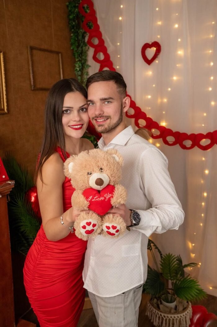 Egy fiatal pár Valentin-napi dekoráció előtt, kezükben egy "I Love You" feliratú plüss macival mosolyognak.