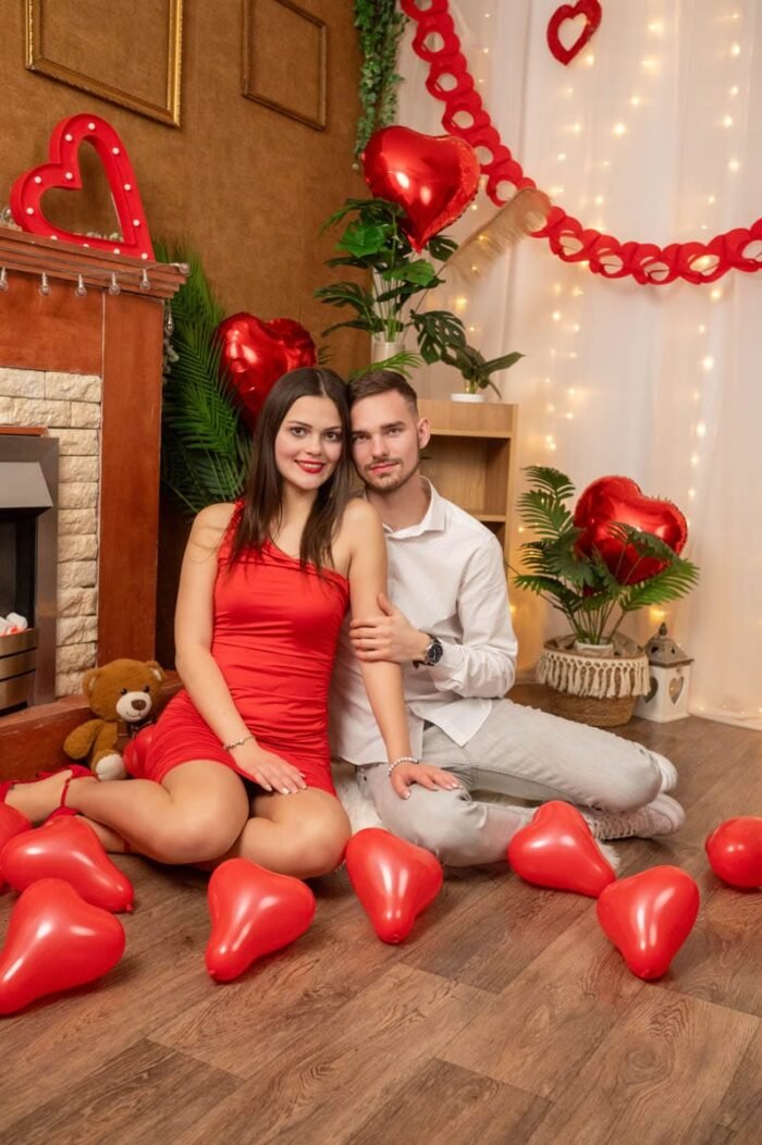 Egy pár Valentin-napi dekorációval körülvéve, a földön ülve, piros lufikkal körülvéve mosolyognak egymás mellett.