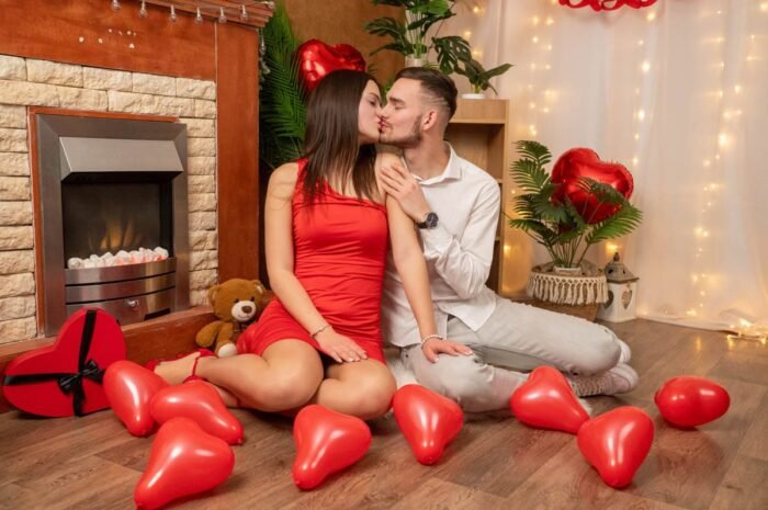 Egy pár Valentin-napi dekorációval körülvéve a földön ül, és megosztanak egy romantikus csókot, piros lufikkal körülvéve, egy kandalló mellett.