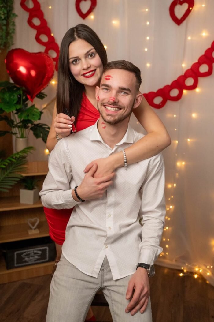 Egy mosolygó férfi csóknyomokkal az arcán, miközben párja vidáman áll mögötte, piros Valentin-napi dekorációval a háttérben.