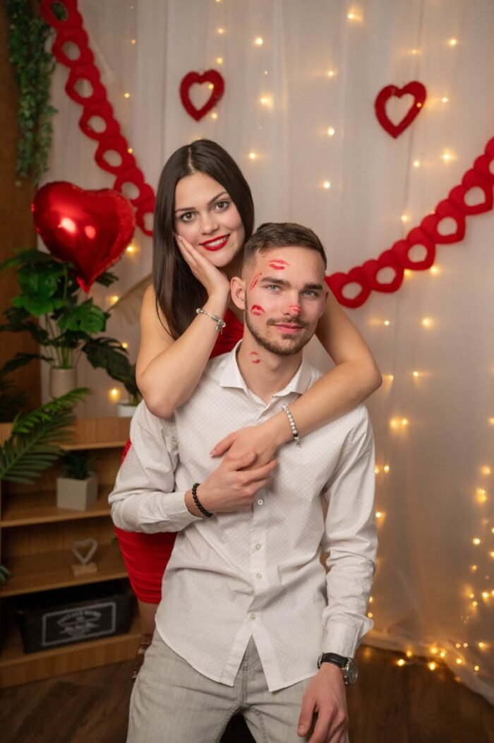 Egy pár Valentin-napi háttér előtt, ahol a férfi arcán csóknyomok láthatók, a nő pedig mosolyogva karolja át hátulról.