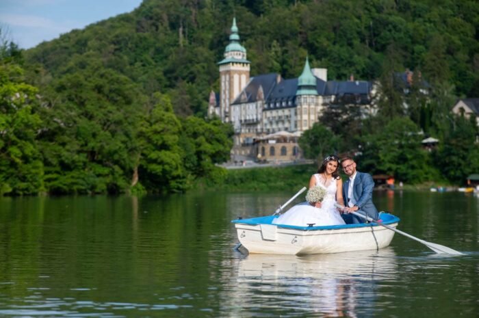 A házasulandó pár csónakban ül a vízen, miközben hátuk mögött a gyönyörű Palotaszálló látható. A férj boldogan mosolyog, míg a feleség virágcsokrot tart, és mindketten élvezik az idilli pillanatot a tó közepén. A zöldellő táj és a napfényes időszak romantikus hangulatot teremt a képen.
