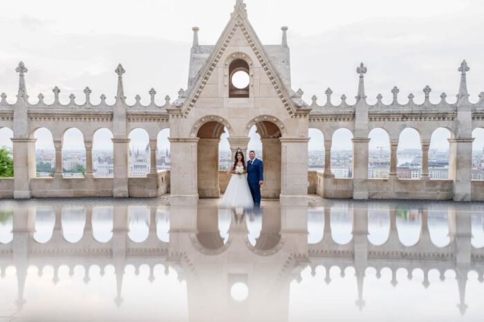 A házaspár egy lenyűgöző pillanatot örökít meg a Halászbástya tetején, miközben a víz tükröződésében a budapesti városkép és a két szerelmes pár vonása egyesül. A táj mögött a Parlament is tisztán látszik, a háttérben pedig a színek gyönyörű játéka.