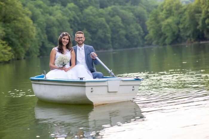 A házasulandó pár egy csónakban ül a vízen, miközben a nyugodt táj és a zöldellő fák veszik körül őket. A feleség virágcsokrot tart, míg a férj, egy evezővel, mellettük ül, mindketten boldogan mosolyognak. A víz tükröződése és a környezet harmóniája kiegészíti a pillanat szépségét.