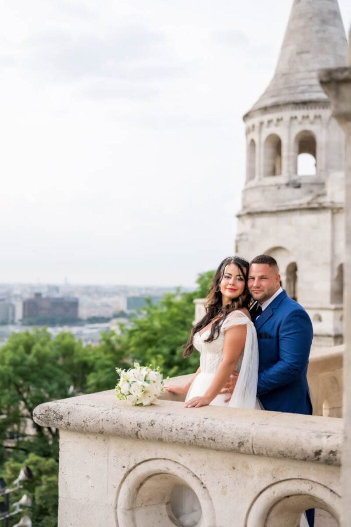 A menyasszony és a vőlegény egy csodálatos pillanatot osztanak meg a Halászbástya kilátóján, miközben Budapest lenyűgöző panorámája terül el előttük. Az arcukon boldog mosoly, háttérben a történelmi város látványa és a szeretet érzése.