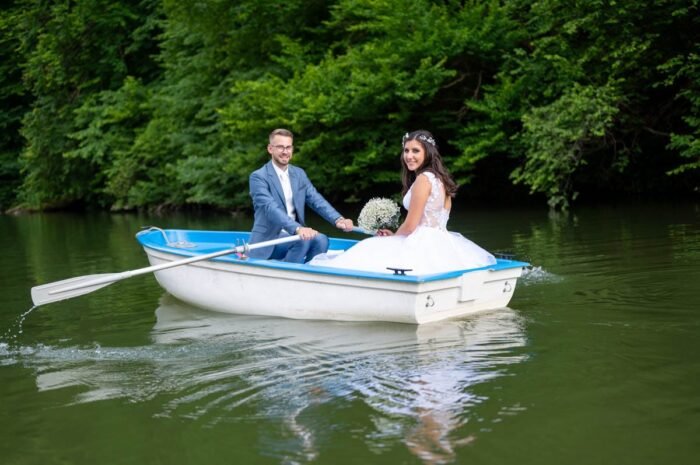 Kreatív esküvői fotós Debrecenben. A fiatal házasok a csónakban, miközben együtt eveznek a nyugodt tavon. A háttérben a zöldellő fák és a víz tükröződése kíséri az ifjú pár boldog pillanatait. A feleség csodálatos menyasszonyi ruhájában, míg férje boldogan irányítja a csónakot, mindketten örömteli pillanatokban osztoznak.