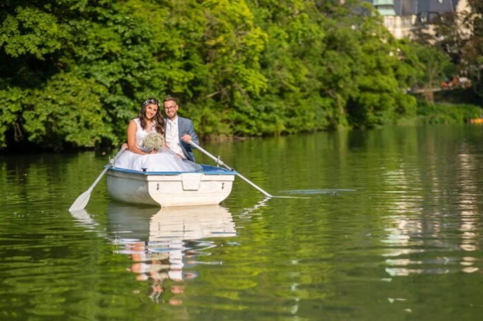 A házaspár boldogan mosolyog egy csónakban, miközben a vízen ringatóznak. A menyasszony gyönyörű fehér esküvői ruhájában és a vőlegény elegáns kék öltönyében ülnek, kezükben a menyasszonyi csokorral. A csónak tükröződése a vízben tökéletesen kiegészíti a romantikus pillanatot, miközben a háttérben zöld növények és egy szálloda épülete látható.