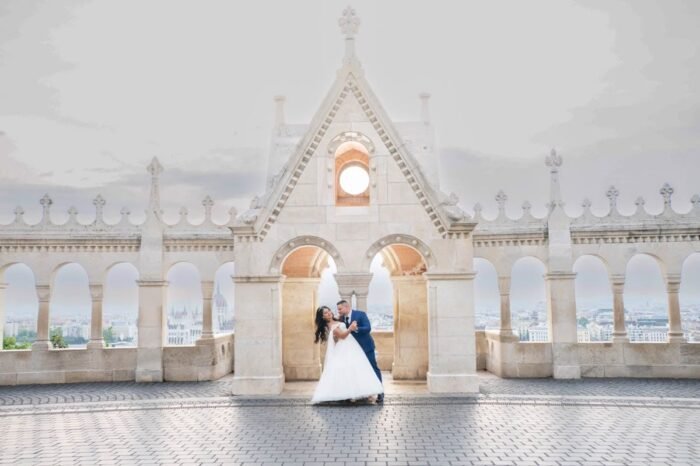 Kreatív fotózás a Budai Várban. A menyasszony és a vőlegény egy boldog pillanatra örökítve a Halászbástya híres pillére alatt, miközben egy romantikus táncot járnak a lenyűgöző Budapest háttérrel. A gyönyörű város látványa és a bástya építészeti részletei teszik különlegessé a pillanatot.