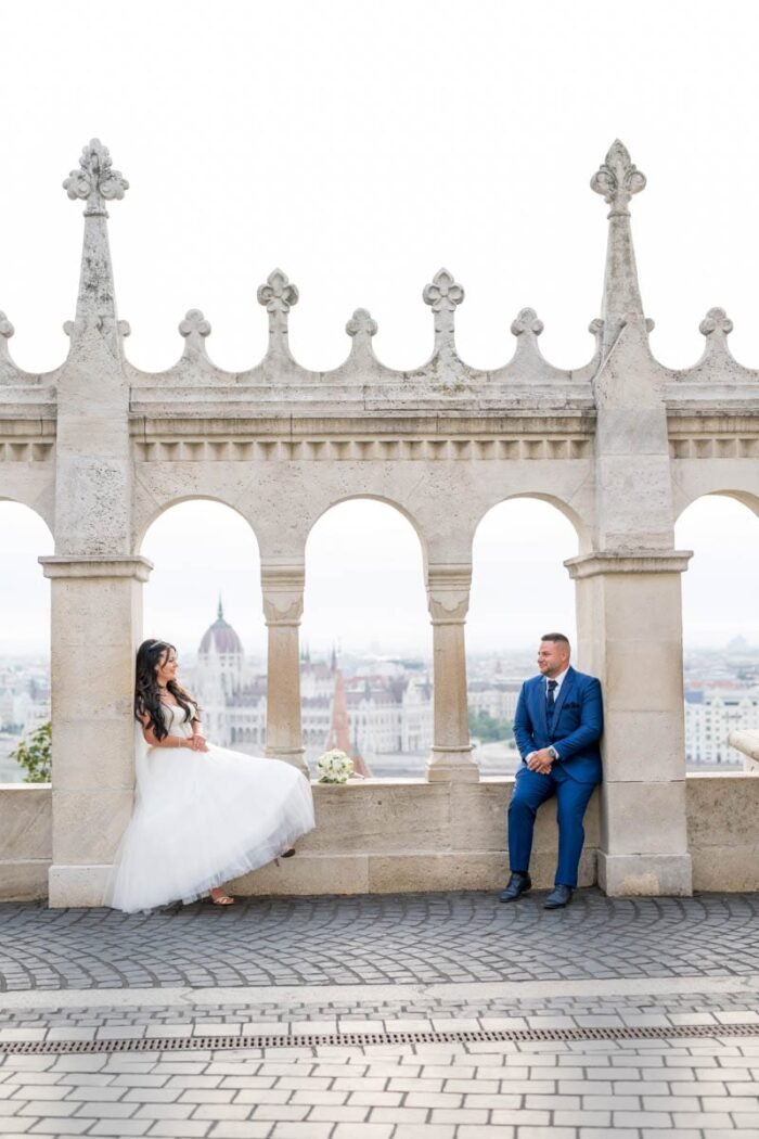 A menyasszony és a vőlegény egy romantikus pillanatra örökítve a Halászbástya teraszán, Budapest csodálatos panorámájával a háttérben. A menyasszony a korláton pihen, miközben mosolyogva nézi a vőlegényt, aki egy kényelmes pozícióban ül a másik oldalon. A Duna, a Parlament és a város látványa teszi a pillanatot még különlegesebbé.