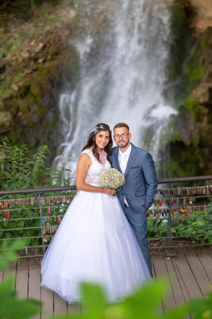 Az ifjú pár boldogan pózol a vízesés előtt, miközben a házasságkötés napján elkapott pillanatokat ünneplik. A háttérben zúgó vízesés és a vízpermet varázslatos hangulata kíséri a boldog házaspár mosolygós fotóját, miközben az összekapcsolt kezeik és a háttérben látható lakatok erősítik a kapcsolat szimbolikus erejét.