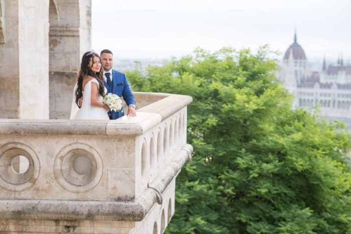 A menyasszony és a vőlegény romantikus pillanatot osztanak meg a Halászbástya teraszán, háttérben Budapest gyönyörű látképével. A menyasszony virágcsokrot tart a kezében, miközben mosolyogva néz a kamerába, a vőlegény mellett, aki öleli őt. A Duna és a Parlament impozáns épületei látszanak a távolban, miközben a természet zöldje és a kőfal finoman keretezi őket.