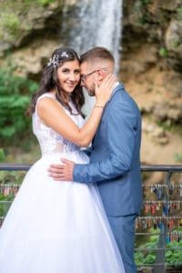 A friss házasok egy romantikus csókot váltanak a vízesés előtt, miközben az esküvői ruha és a kék öltöny harmonikus kontrasztot alkot a háttérben található vízeséssel. A házaspár boldog pillanata a szeretet és intimitás érzését sugározza, miközben a vízesés zúgása és a lakatokat tartó híd teszi még varázslatosabbá a környezetet.