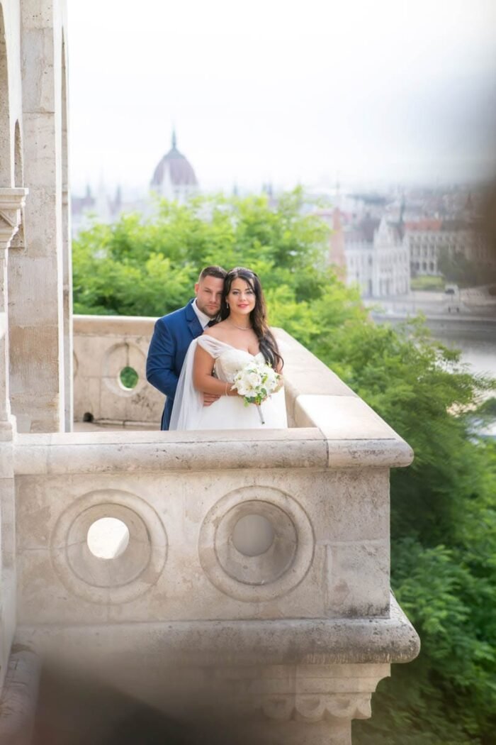 Az ifjú pár egy romantikus pillanatot oszt meg a Halászbástya teraszán, a menyasszony gyönyörű fehér ruhájában és virágcsokorral a kezében. A vőlegény hátulról öleli át feleségét, miközben a háttérben Budapest impozáns látképe és a Duna látható. A napfény lágyan megvilágítja őket, hangsúlyozva kapcsolatuk intimitását és boldogságát.
