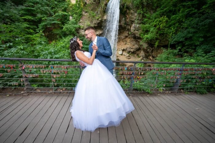 A friss házasok egy hídon táncolnak, miközben a háttérben egy festői vízesés és egy padlóval borított híd látható. A híd korlátján lakatokat helyeztek el, amelyek szerelmi szimbólumokat jelképeznek. A pár boldogan egymásra néz, miközben a menyasszony hosszú fehér esküvői ruhája elragadóan kiemelkedik a természetes tájból.