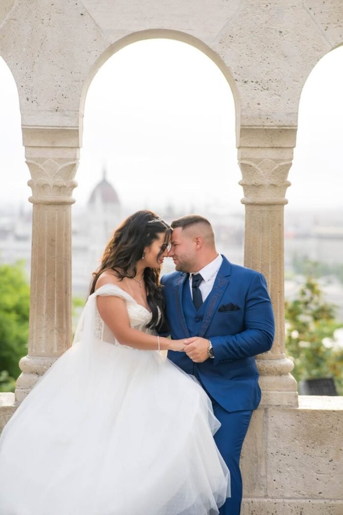 Budapesti esküvői fotós. Az ifjú pár egymásra hajolva, kézen fogva áll a Halászbástya egyik ablakában, háttérben Budapest impozáns látképével. A menyasszony fehér esküvői ruhájában és tiarájával, míg a vőlegény elegáns kék öltönyében ragyogó pillanatokban osztozik feleségével. A napfény lágy fényekkel öleli körbe őket, miközben szoros kapcsolatuk tükröződik az arcaikon.