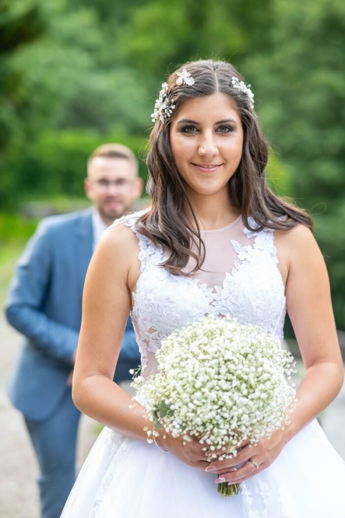 A menyasszony a gyönyörű fehér ruhájában, kezében egy nagy csokor gyöngyvirággal, mosolyogva áll a háttérben, ahol a vőlegény részben homályosan látszik. A háttérben zöld növények és egy sétaút alkotják a természetes környezetet.