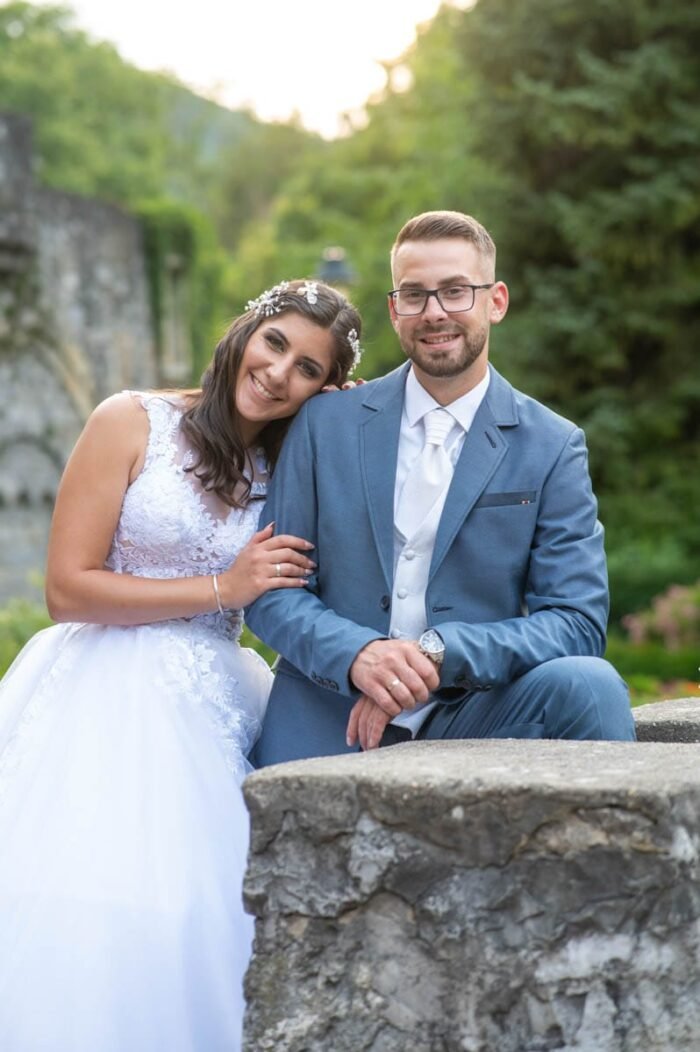 A menyasszony és a vőlegény egy kőkerítésnél ülnek, mosolyogva egymásra néznek. A menyasszony gyönyörű fehér ruhája tökéletesen illeszkedik a természetes környezethez, míg a vőlegény elegáns kék öltönyében boldogan tartja a felesége kezét. A háttérben zöldellő fák és virágos kertek adják a festői tájat.