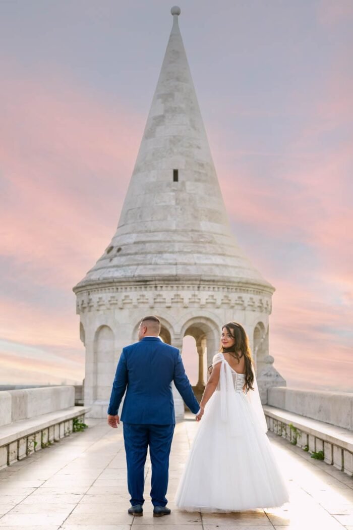 A házaspár kézen fogva sétál a Halászbástya teraszán, Budapest városának lenyűgöző látképével a háttérben. A menyasszony fehér esküvői ruhájában, míg a vőlegény elegáns kék öltönyben van. A háttérben a bástya jellegzetes tornya és a színes naplemente látható.