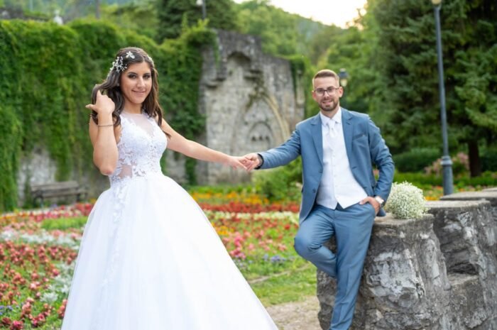 Esküvői fotózás Debrecenben. A menyasszony és vőlegény egy virágos kertben sétálnak, miközben kezüket fogják. A menyasszony gyönyörű fehér esküvői ruhájában mosolyog, a vőlegény kék öltönyében boldogan áll mellette, a háttérben a természet zöldell és a virágok színes kavalkádot alkotnak.