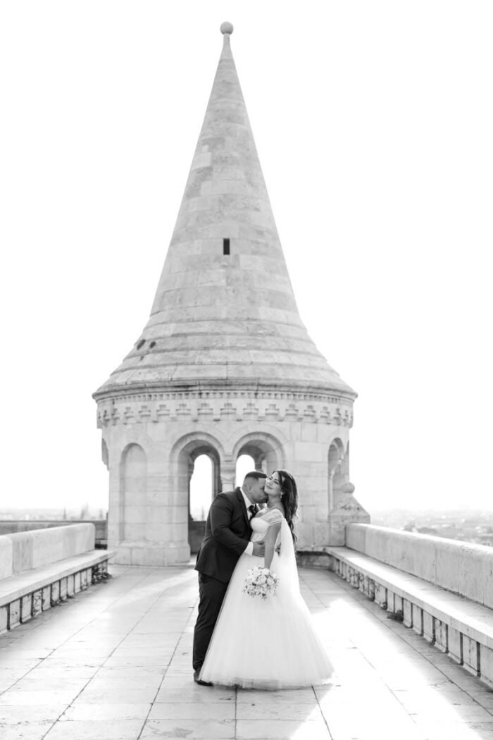 A fiatal pár a Halászbástya teraszán, Budapest városára nézve osztozik egy meghitt csókban. A menyasszony gyönyörű fehér esküvői ruhájában, virágcsokrot tartva, míg a vőlegény fekete öltönyében öleli át őt. A háttérben a bástya jellegzetes tornya és a fényes városi látkép látható.