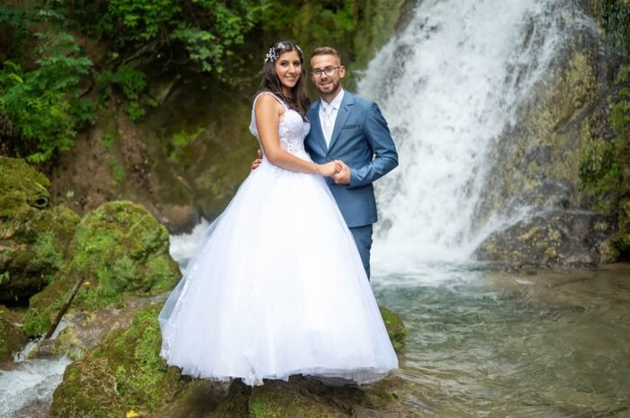 A boldog pár összefonja kezeit a vízesés előtt, miközben a természet gyönyörű háttérként szolgál számukra. A menyasszony fehér ruhája ragyog a vízesés frissítő környezetében, a férj pedig elegáns kék öltönyében mosolyog.