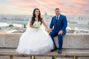 A fiatal pár a Budai Várnál ül a falon, kézen fogva, miközben a város csodálatos panorámáját és a Parlament épületét láthatják a háttérben. A menyasszony fehér esküvői ruhát visel, virágcsokrot tart a kezében, míg a vőlegény kék öltönyt visel. A naplemente színei meleg fényt adnak a romantikus pillanathoz.
