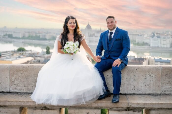 A fiatal pár a Budai Várnál ül a falon, kézen fogva, miközben a város csodálatos panorámáját és a Parlament épületét láthatják a háttérben. A menyasszony fehér esküvői ruhát visel, virágcsokrot tart a kezében, míg a vőlegény kék öltönyt visel. A naplemente színei meleg fényt adnak a romantikus pillanathoz.