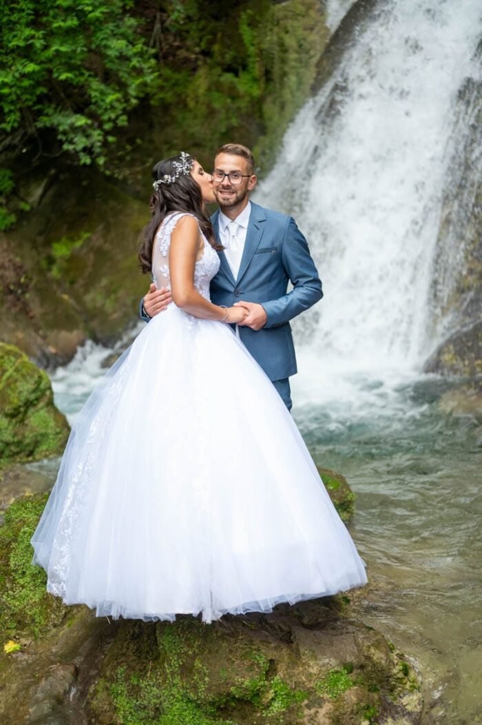 A pár csókot vált a vízesés előtti sziklán, miközben körülöttük zöldellő növények és friss víz folyik. Az esküvői ruha és a kék öltöny lenyűgöző kontrasztot alkot a természet csodáival.