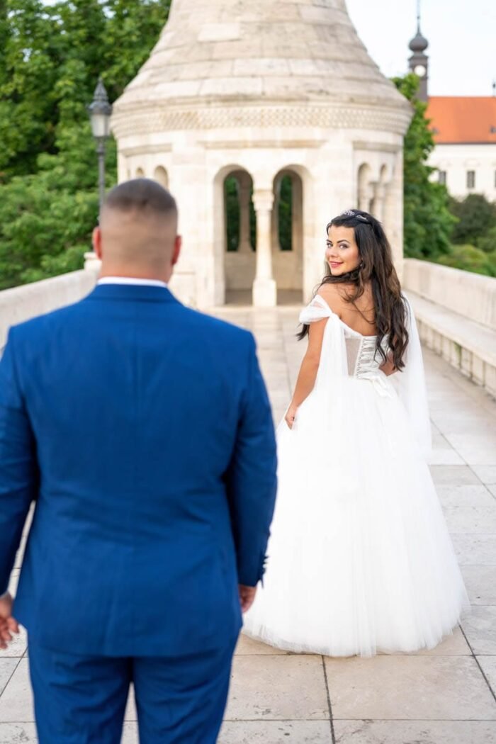 A menyasszony visszanéz mosolyogva, miközben az ifjú férje, aki kék öltönyben van, mögötte sétál. A háttérben a Halászbástya egyik toronyrésze látszik, és a környezet zöld fákban gazdag, mely romantikus hangulatot áraszt.