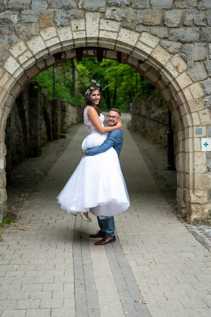 Esküvői kreatív fotózás Lillafüreden A férj felemeli feleségét egy romantikus sétán, miközben mosolyognak a fényképezőgépbe. A háttérben egy gyönyörű kőhíd ível át az úton, amely a varázslatos hangulatot erősíti a pillanaton.