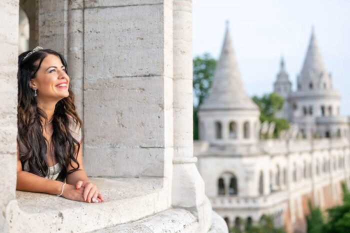 A menyasszony boldogan néz a távolba, miközben a Budai Vár környezetében található Halászbástyán áll. A háttérben a bástya jellegzetes toronyrészei látszanak, miközben a menyasszony gyönyörű mosollyal élvezi a pillanatot.