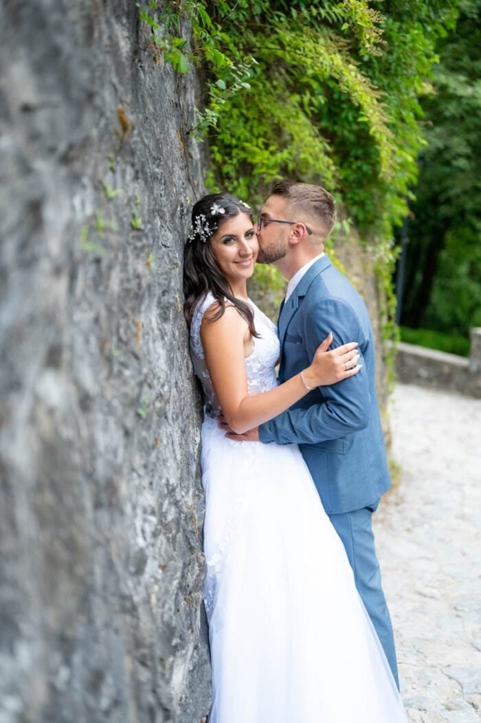 A vőlegény kedvesen megcsókolja a menyasszonyt, miközben ő mosolyogva néz a fényképezőgépbe. A háttérben a kőfal és a zöld növények a romantikus hangulatot erősítik.