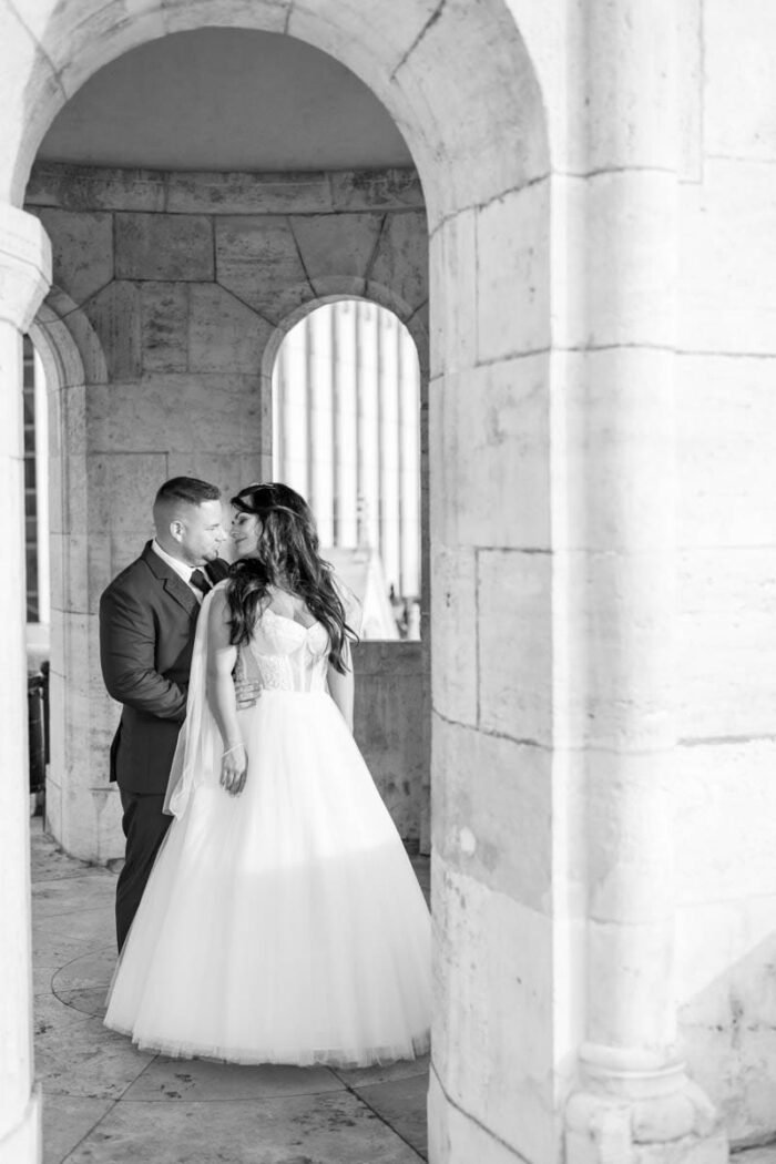 A friss házasok romantikus pillanata a Budai Vár egyik történelmi helyszínén. A pár a vár falai között áll, egymásra nézve, miközben a háttérben a város egy darabja is látszik. A kép fekete-fehér, kiemelve a házaspár érzelmes összhangját.
