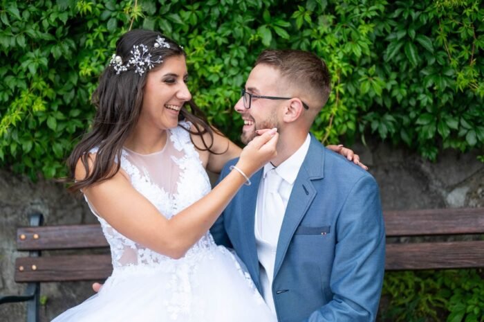 A menyasszony gyengéden simítja a vőlegény arcát, miközben mindketten boldogan nevetnek a háttérben a zöld növények előtt. A menyasszony fehér ruhája és a vőlegény szürke öltönyje tökéletesen illenek egymáshoz.