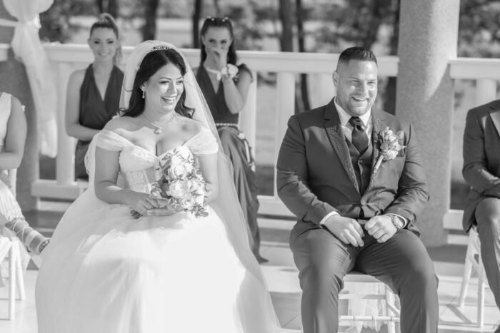 A boldog ifjú pár az esküvői szertartás alatt ül, miközben mindketten nevetnek és egymásra néznek. A háttérben az esküvő vendégei is figyelik őket, a kép fekete-fehérben készült.