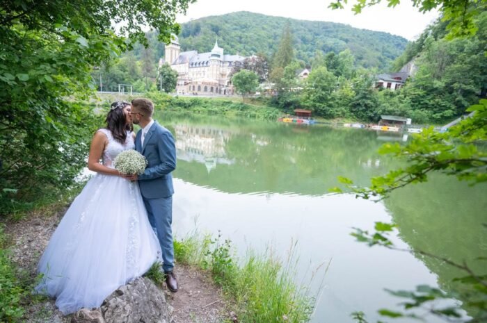 A friss házasok csókot váltanak a tóparton, miközben a gyönyörű menyasszony fehér ruhája csillog a víz tükrében. A háttérben a szálloda és a hegyek látszanak, miközben a környező természet zöldjével öleli körbe őket.