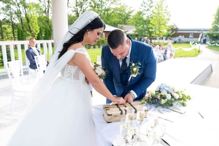 Az ifjú pár éppen a ceremónia alatt egy szertartásos dobozt zár le, amely a boldogságot szimbolizálja. Az alapszín fehér, a háttérben a vendégek figyelemmel kísérik az eseményt.