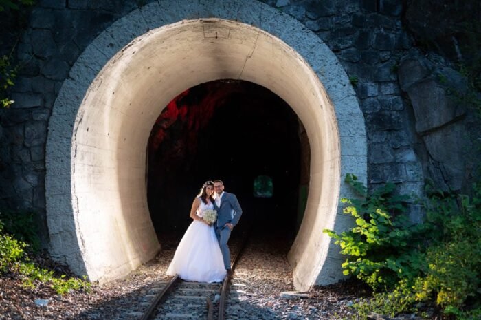 A boldog pár egy vasúti alagút előtt pózol, miközben a háttérben vörös fények világítanak, és a vasúti sínek vezetnek az alagút belsejébe. A menyasszony gyönyörű fehér esküvői ruhájában, a vőlegény pedig elegáns kék öltönyében öleli őt, és a romantikus pillanatok a varázslatos helyszíntől függetlenül ragyognak.