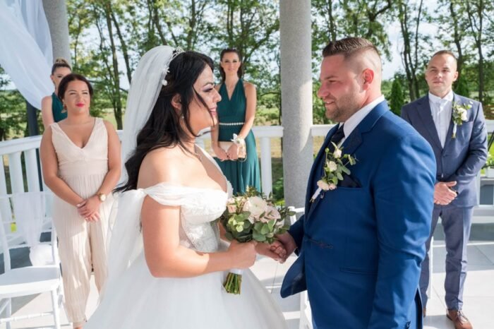 Az ifjú pár egymásra nézve mondja el esküvői fogadalmát, miközben boldogan tartják egymás kezét. A háttérben az esküvői szertartás tanúi és a vendégek figyelik a meghitt pillanatot.