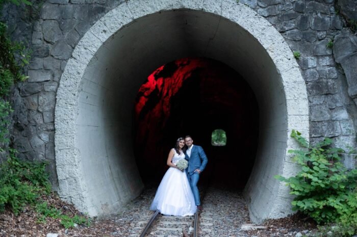 A friss házasok boldogan pózolnak egy vasúti alagút előtt, miközben a háttérben titokzatos, vörösen megvilágított alagúton átvilágít a nap. A menyasszony fehér esküvői ruhában és a vőlegény kék öltönyében ölelik egymást, miközben a vasúti sínekre álltak, így az alagút lenyűgöző formája és a színvilág remekül kiegészíti a romantikus pillanatot