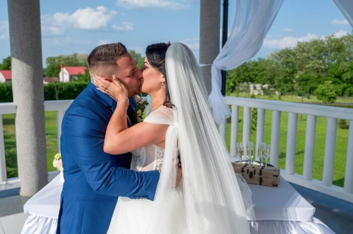 Az ifjú pár boldogan csókolózik az esküvői szertartás alatt, miközben a háttérben a friss házasok körüli vendégek figyelik a pillanatot. Az öröm és szeretet érzése látható a boldog pár arcán.