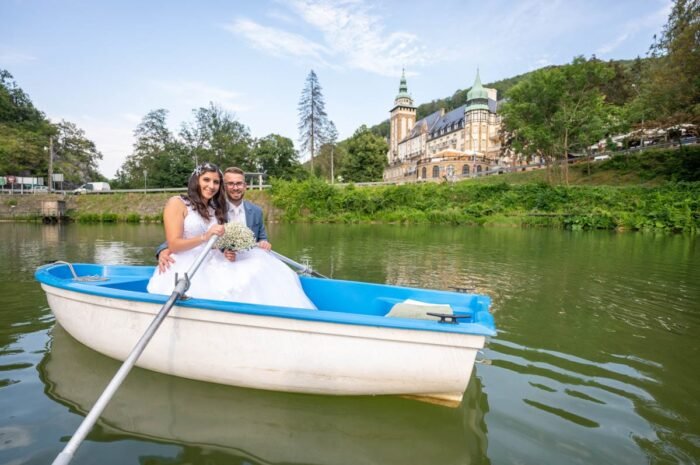 A fiatal házaspár boldogan mosolyog egy csónakban, miközben a vízen ringatóznak. A menyasszony gyönyörű fehér esküvői ruhájában és a vőlegény elegáns kék öltönyében ülnek. A háttérben a híres Palotaszálló impozáns épülete emelkedik, zöldellő természet környezetében. A csónak tükröződése a vízen tökéletesen kiegészíti a romantikus pillanatot.