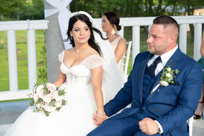 A friss házasok kéz a kézben ülnek a szertartás alatt, miközben az ifjú feleség tekintete a jövőbe, férje pedig az eseményre koncentrál. A háttérben a szertartás további résztvevői láthatók, míg a boldog pár életük legfontosabb pillanatait éli át.