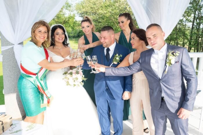 Az ifjú pár és közeli barátaik egy koccintás közepette ünneplik a boldog pillanatokat, miközben a szertartás helyszíne mögött zöldellő táj és baráti társaság övezi őket. Az esemény örömteli hangulatát a boldog mosolygó vendégek és a friss házasok fokozzák.