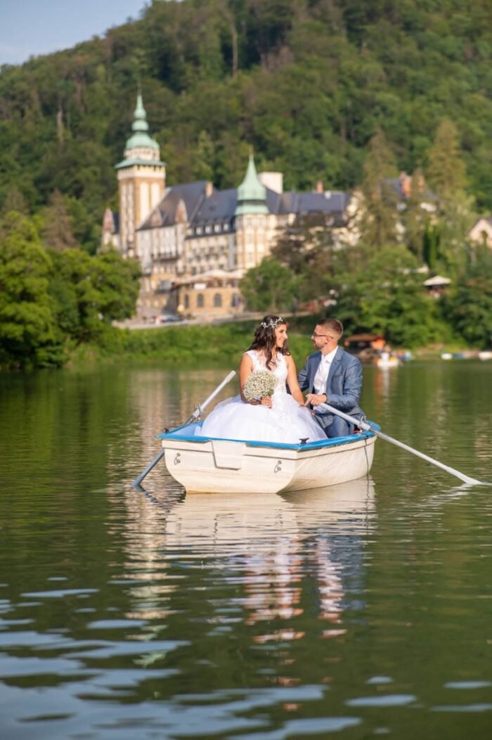 Esküvői fotós hajdúböszörmény - A friss házasok egy kis csónakban ülnek, miközben egymás szemébe néznek. A menyasszony gyönyörű esküvői ruhájában, míg a vőlegény elegáns kék öltönyben látható. A csónak a tó közepén ringatózik, a háttérben a Palotaszálló impozáns épülete emelkedik. A vízen való tükröződés gyönyörű kiegészítője a romantikus pillanatnak.