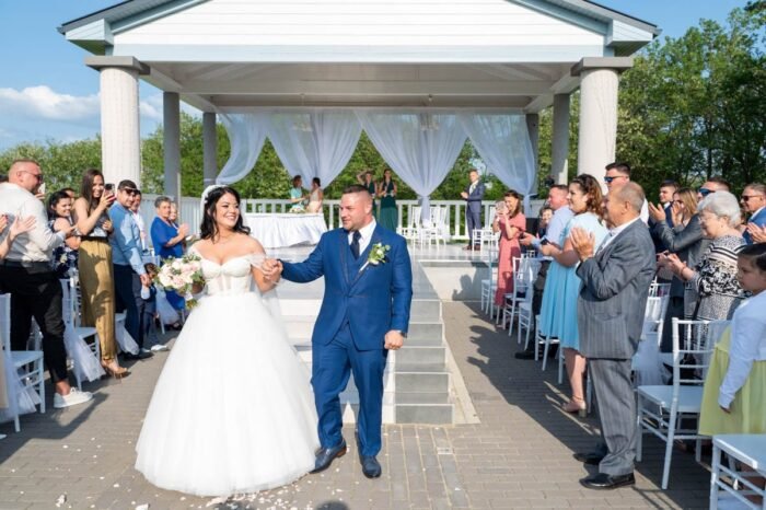 Az ifjú pár boldogan lépked a házasságkötés után, miközben vendégeik tapsolnak és ünneplik őket. A friss házasok mosolyogva, kéz a kézben vonulnak, miközben a napfényben virágok hullanak körülöttük. Az esküvői szertartás meghitt pillanatai örökre megmaradnak.