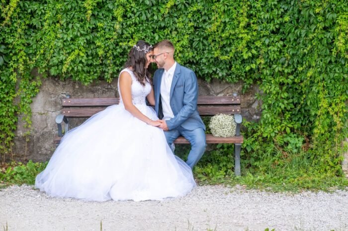 A fiatal pár egy padon ülve, boldogan néznek egymás szemébe. A menyasszony gyönyörű fehér esküvői ruhát visel, míg a vőlegény elegáns kék öltönyben.Esküvői videó készítés Debrecenben